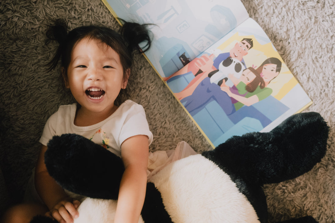 A joyful toddler lying on a carpet next to a stuffed panda, laughing while reading a bilingual children’s book with an illustration of a family holding a stuffed panda, creating a warm and playful moment.