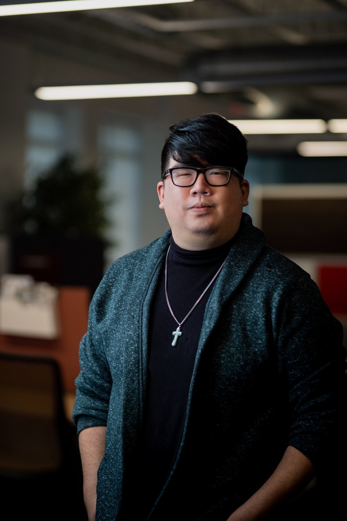 An Asian man wearing glasses, a dark sweater, and a cross necklace, standing confidently in a modern office setting with blurred background elements of desks and greenery