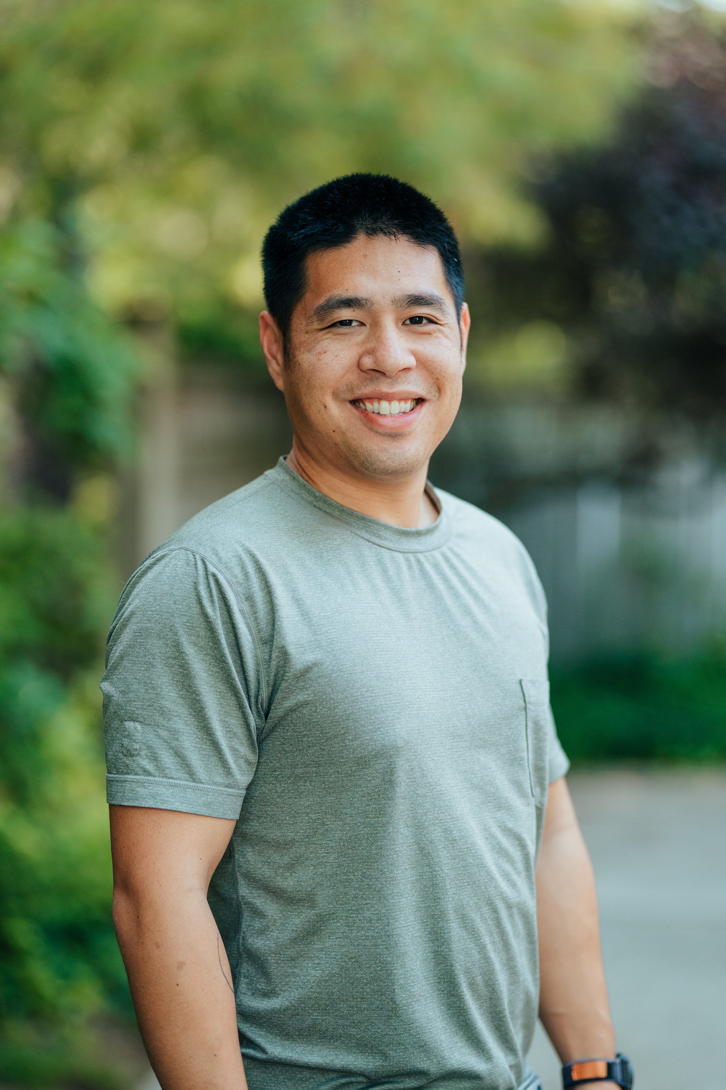 A smiling Asian man wearing a green shirt, standing outdoors with a blurred natural background of greenery and trees, conveying a warm and approachable demeanor.