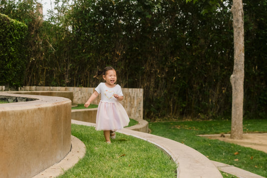 A joyful toddler in a white and pink dress running outdoors on a curved pathway, surrounded by green grass and trees.