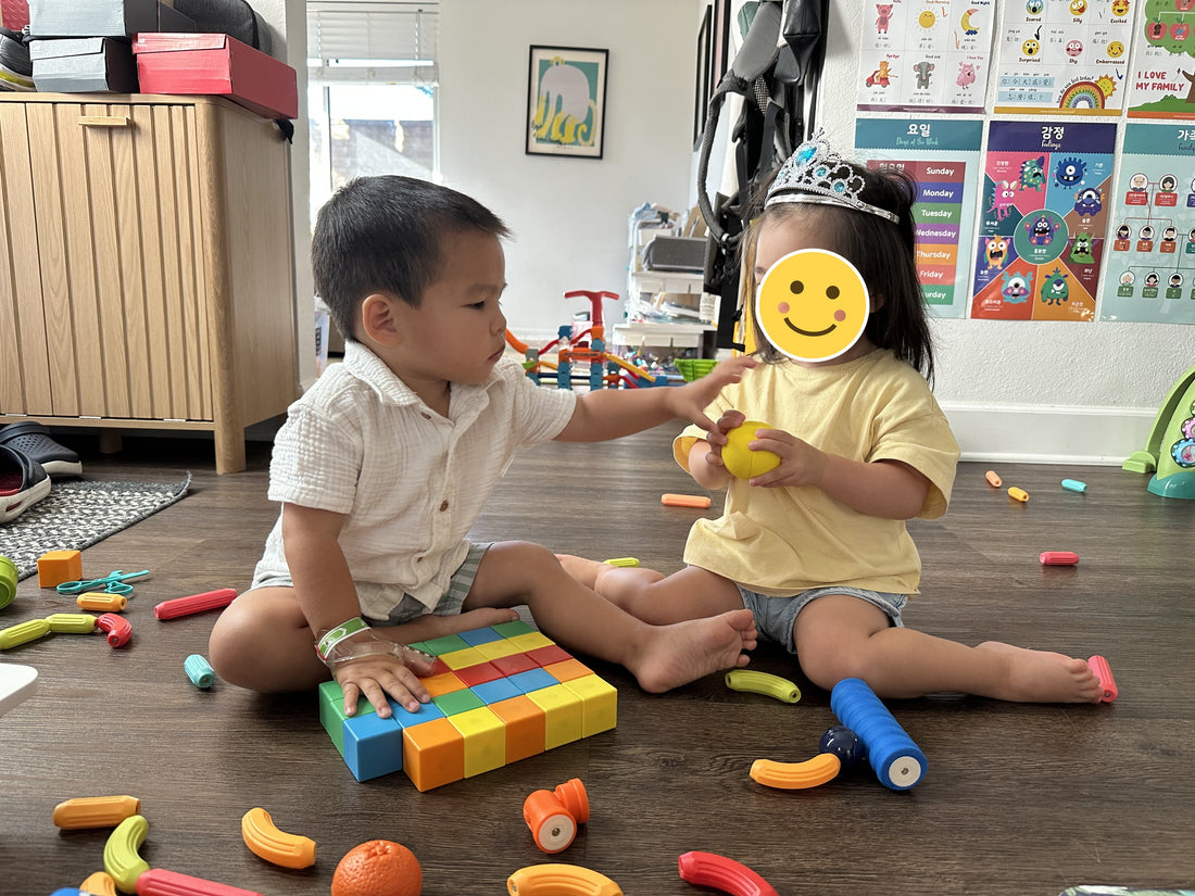 Two toddlers playing with toys on the floor, one reaching toward a ball, the other's face covered by a yellow smiley sticker