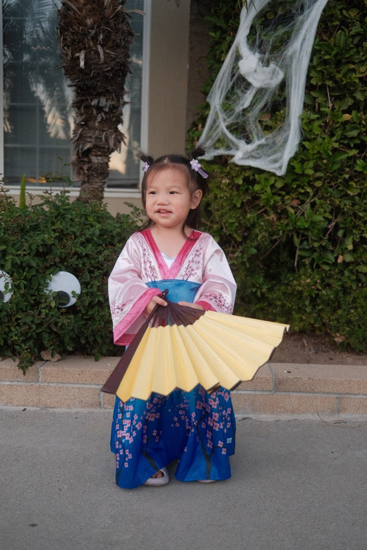 Toddler in a cherry-blossom kimono Halloween costume holding a folding fan, with spider-web decorations behind her