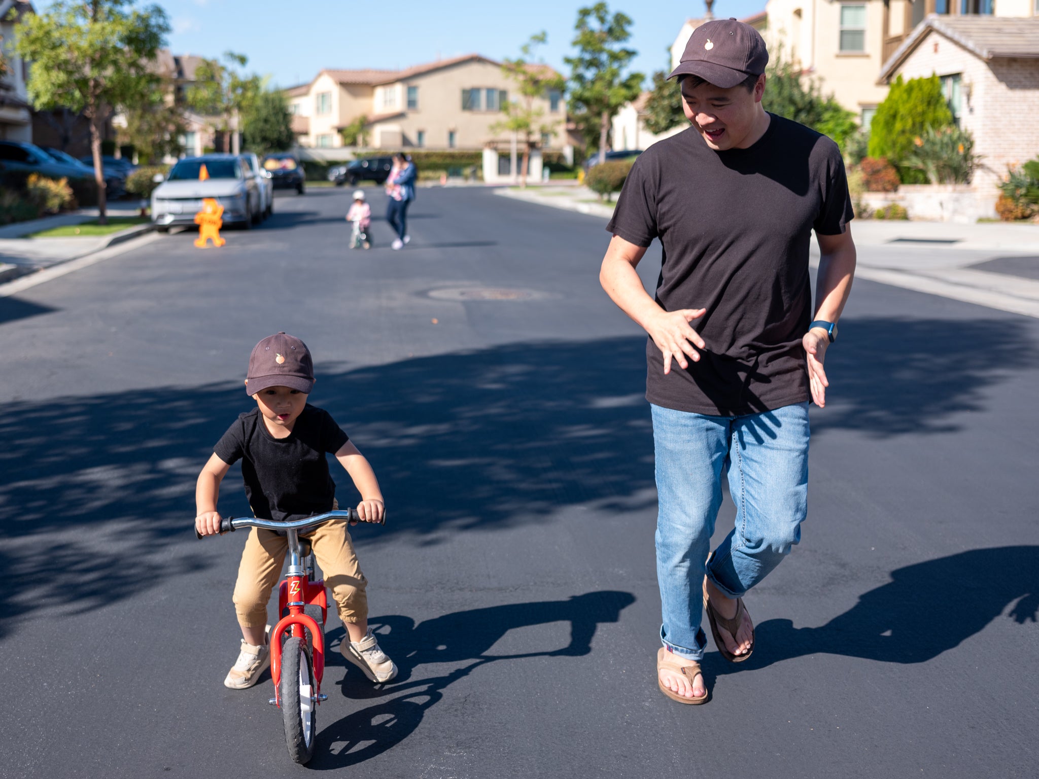 A father and son wearing matching hats and black shirts, enjoying a sunny day outdoors as the child rides a red balance bike while the father walks alongside, encouraging him in a suburban neighborhood setting.