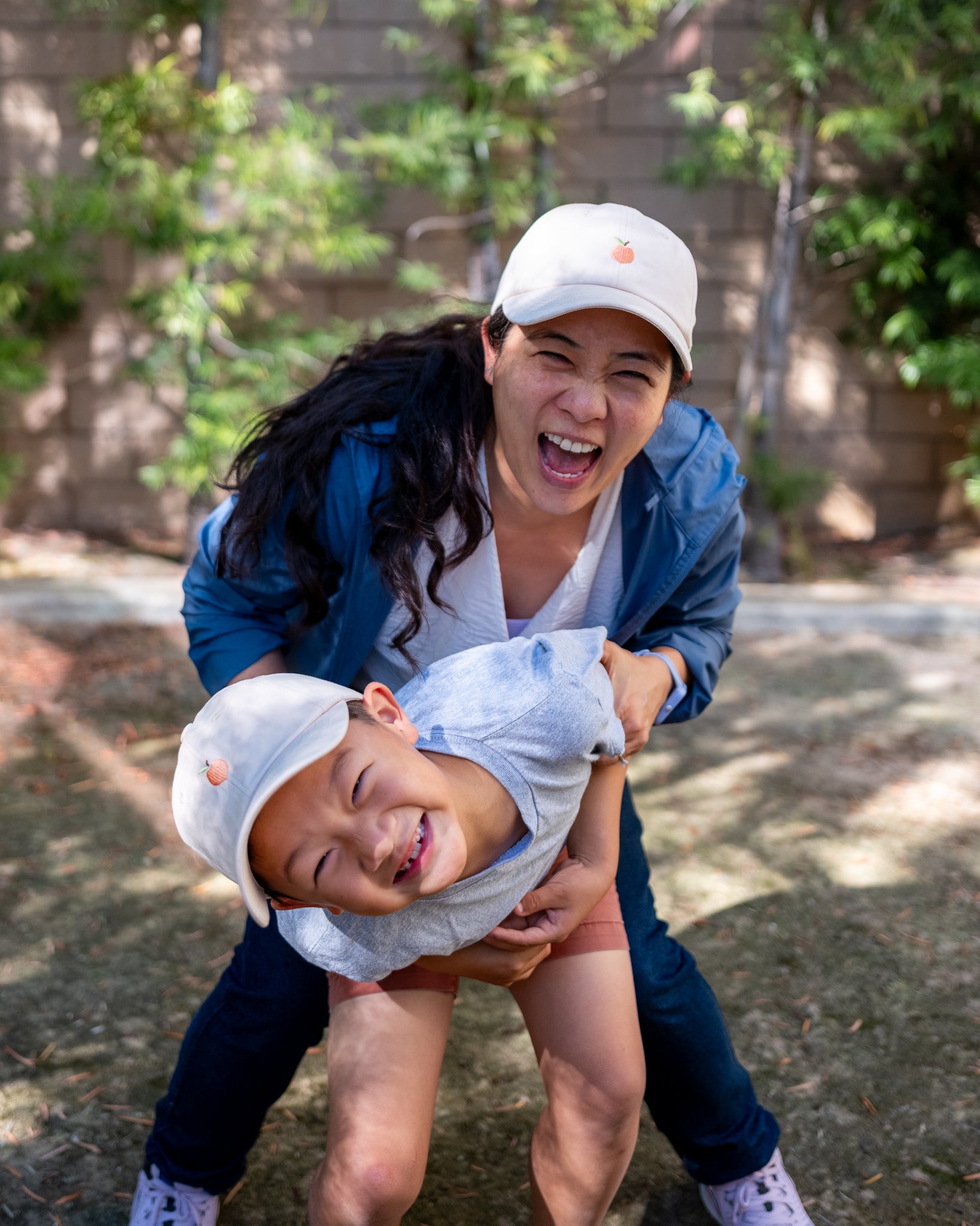 A joyful mother and son wearing matching beige hats with a peach logo, laughing together outdoors on a sunny day, surrounded by greenery and creating a playful and loving moment.