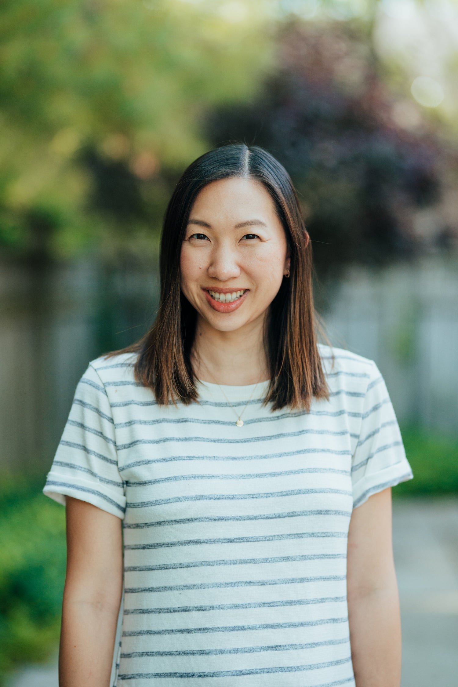 A smiling Asian woman wearing a striped shirt and a delicate necklace, standing outdoors with a blurred background of greenery and trees, radiating warmth and friendliness.